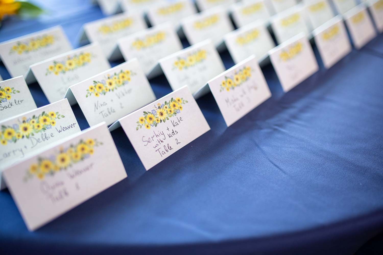Sunflower-decorated wedding place cards arranged on blue tablecloth for reception seating.