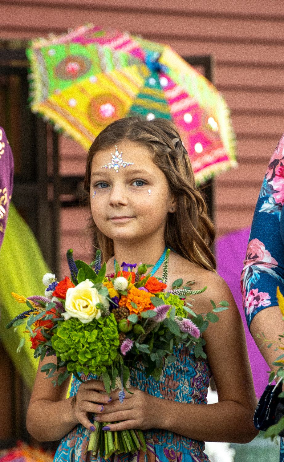 Young flower girl in blue floral dress holds vibrant bouquet with jewel forehead decoration at festive outdoor wedding.