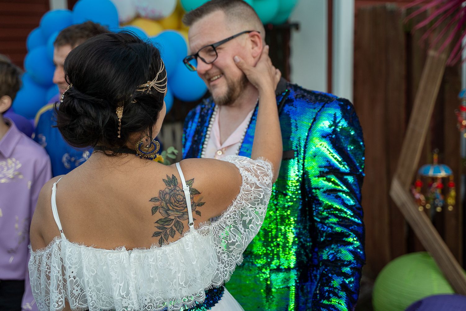 Bride and groom share a tender dance moment, bride showing rose tattoo, groom in green sequin jacket at colorful wedding.