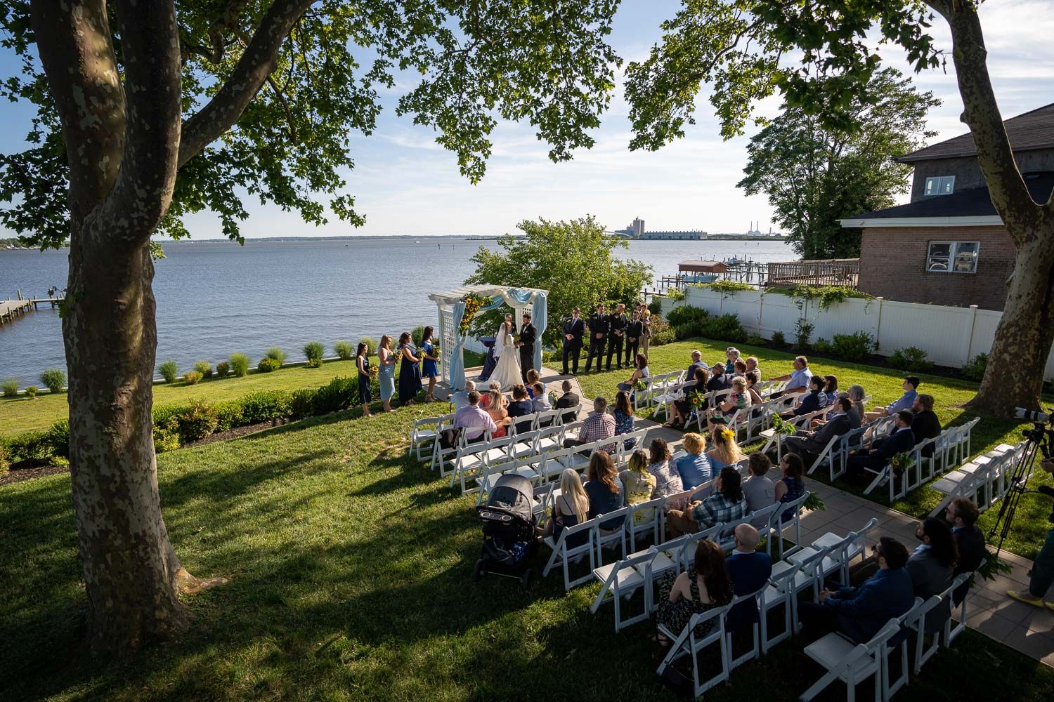 Aerial view of intimate outdoor waterfront wedding ceremony with white chairs and guests on green lawn.