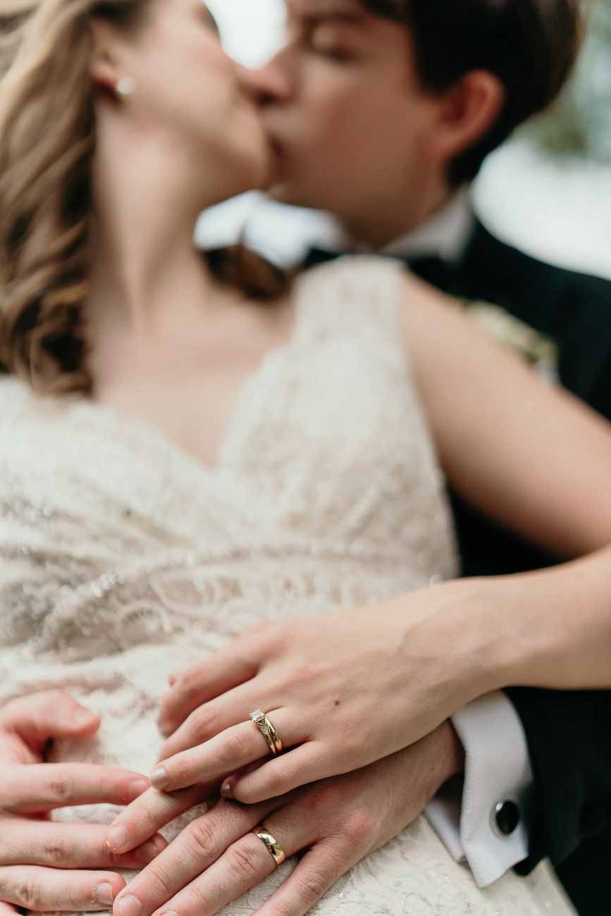 Close-up of newlyweds kissing while displaying their gold wedding rings on clasped hands.