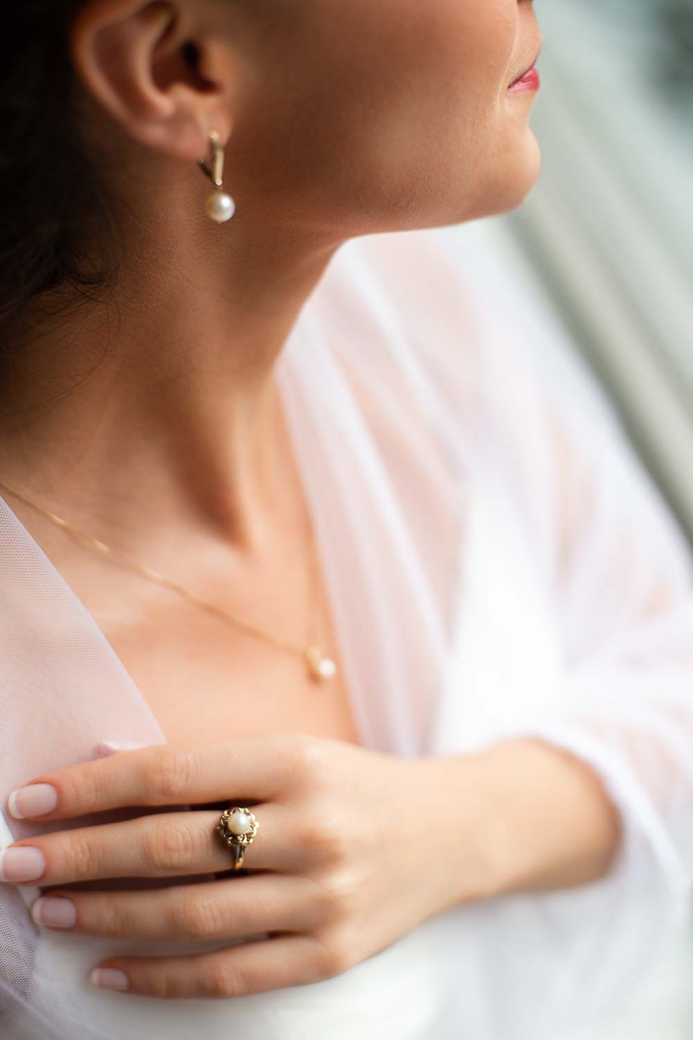 Close-up of bride wearing elegant pearl drop earring, gold ring, and delicate necklace in soft light.