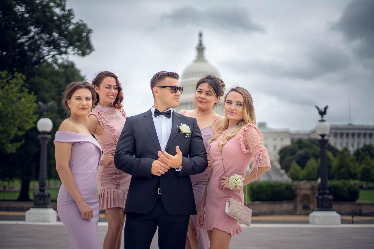 Groom in tuxedo poses with four bridesmaids in pink dresses near US Capitol building.