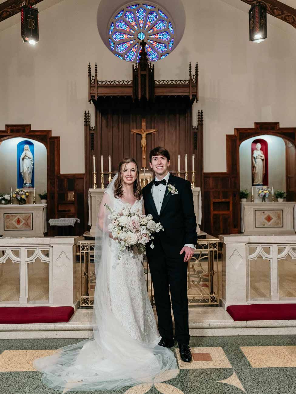 Bride in lace gown and groom in tuxedo pose at Catholic church altar after wedding ceremony.
