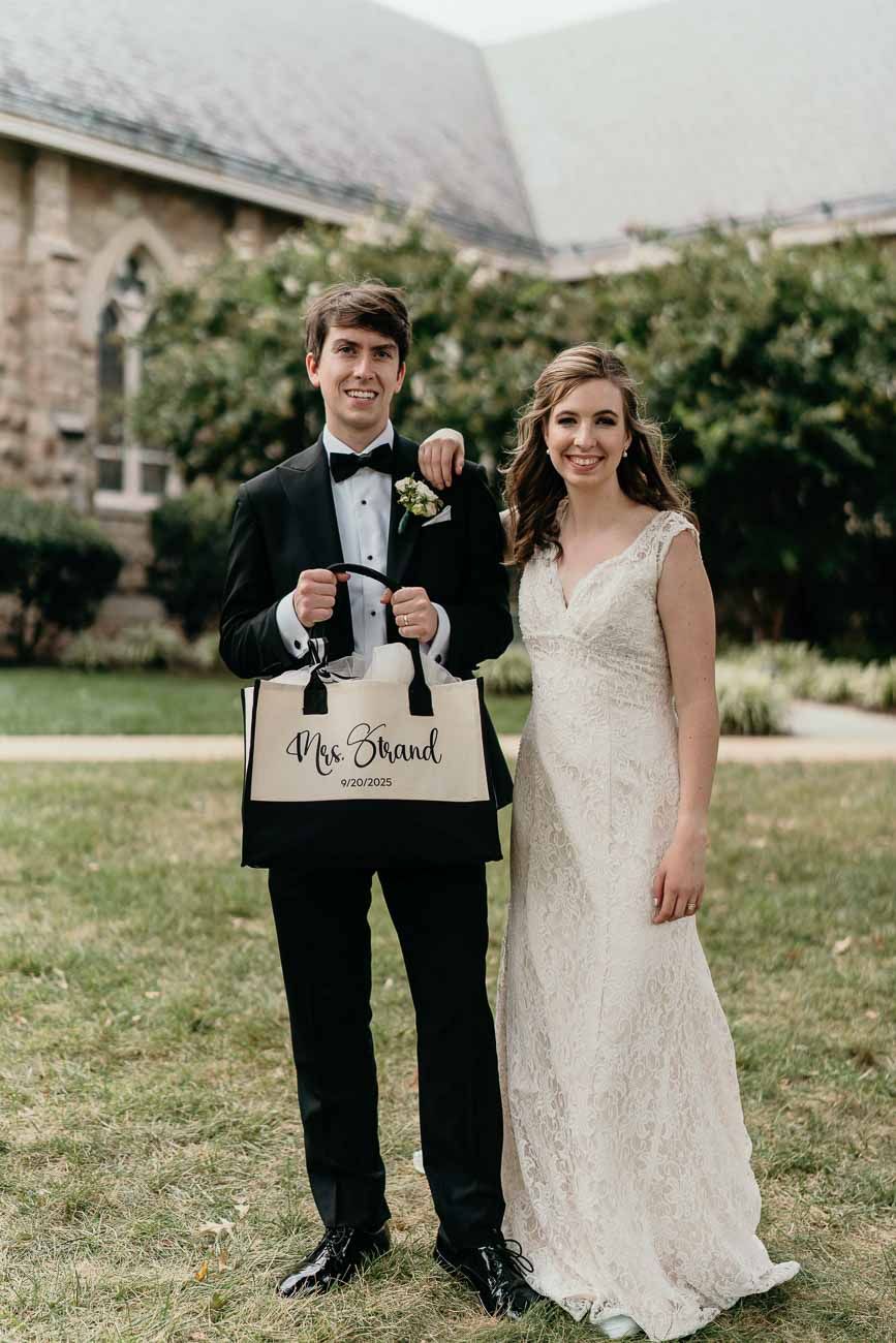 Newlyweds pose together holding a Mrs. Brand tote bag outside a stone church after their wedding.