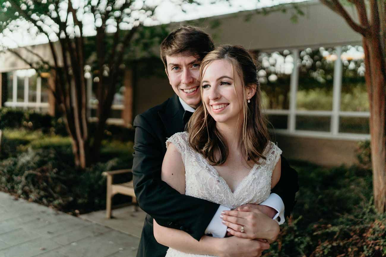 Groom hugging bride from behind as both smile warmly in an outdoor garden setting.