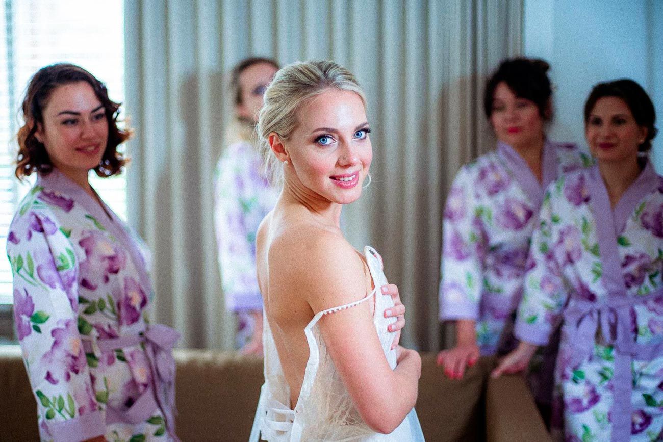 Blonde bride in white wedding dress smiling over shoulder with bridesmaids in floral robes behind.