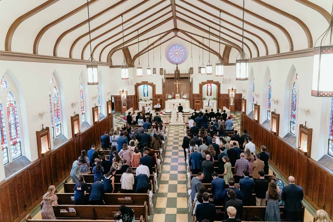 Overhead view of Catholic church interior with wedding guests seated in wooden pews during ceremony.