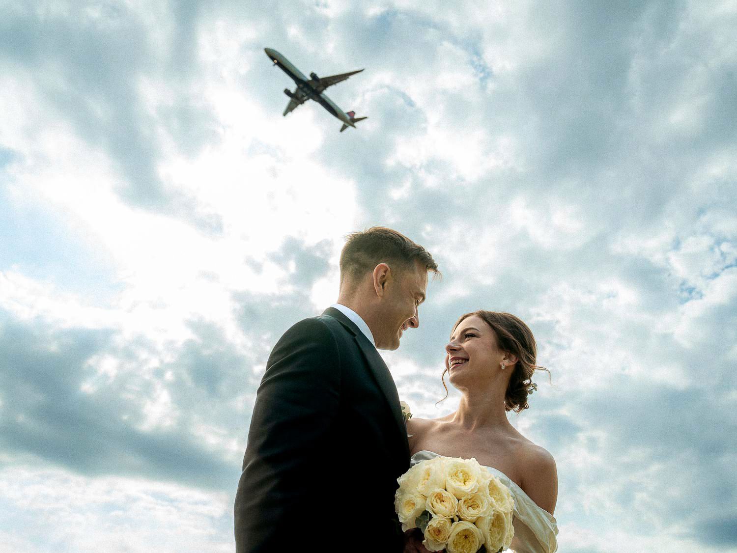 Smiling bride and groom look at each other with airplane flying overhead against dramatic cloudy sky.