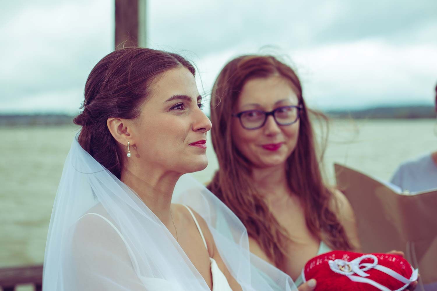 Bride in white veil smiles during ceremony as bridesmaid with red glasses stands beside her outdoors.