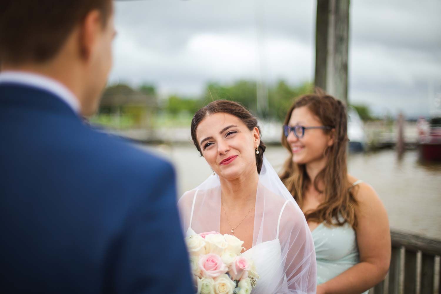 Radiant bride smiles lovingly at groom while holding pink and white bouquet with bridesmaid beside her.