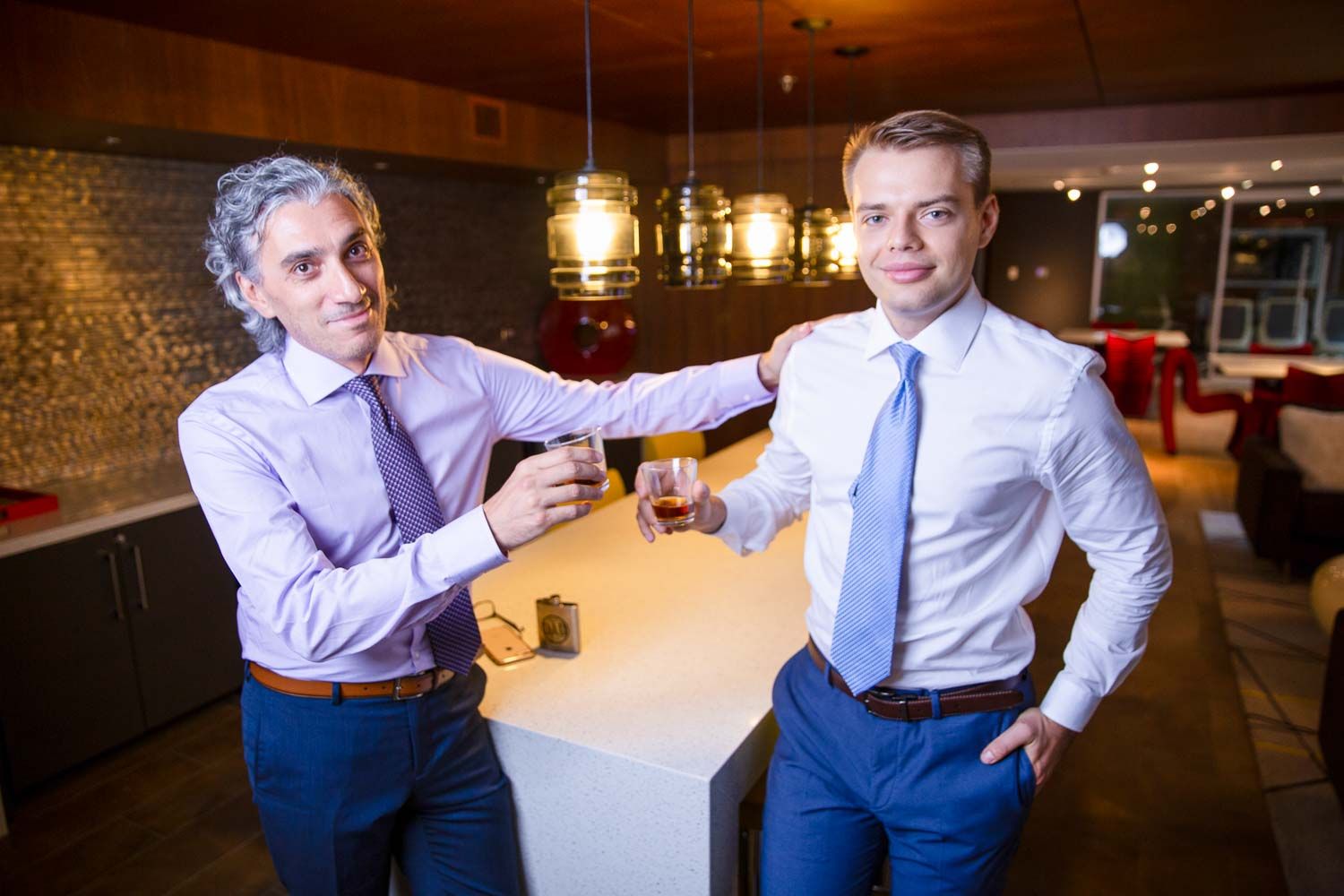 Two men in suits toasting drinks at a modern bar, smiling in a warmly lit restaurant setting.