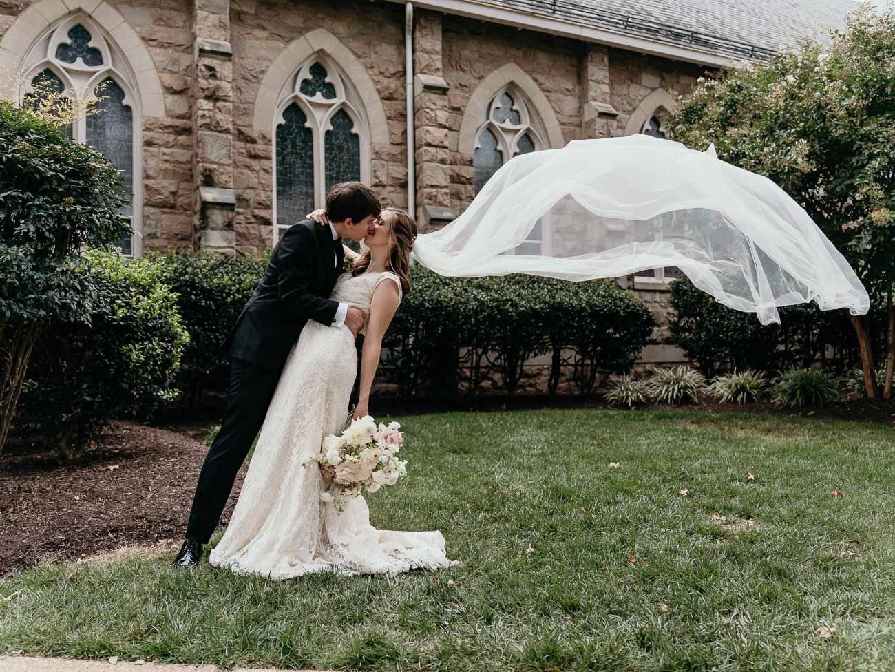 Groom dips bride for a kiss outside stone church as her veil dramatically billows in the wind.