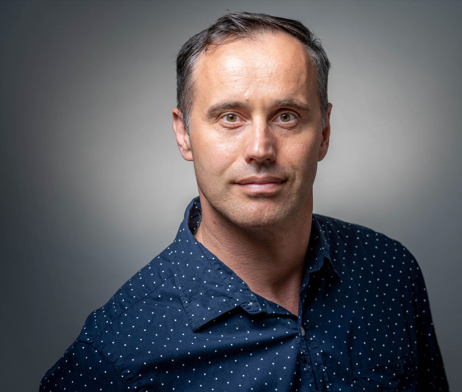 Professional headshot of a middle-aged man in a navy polka dot shirt against a gray background.