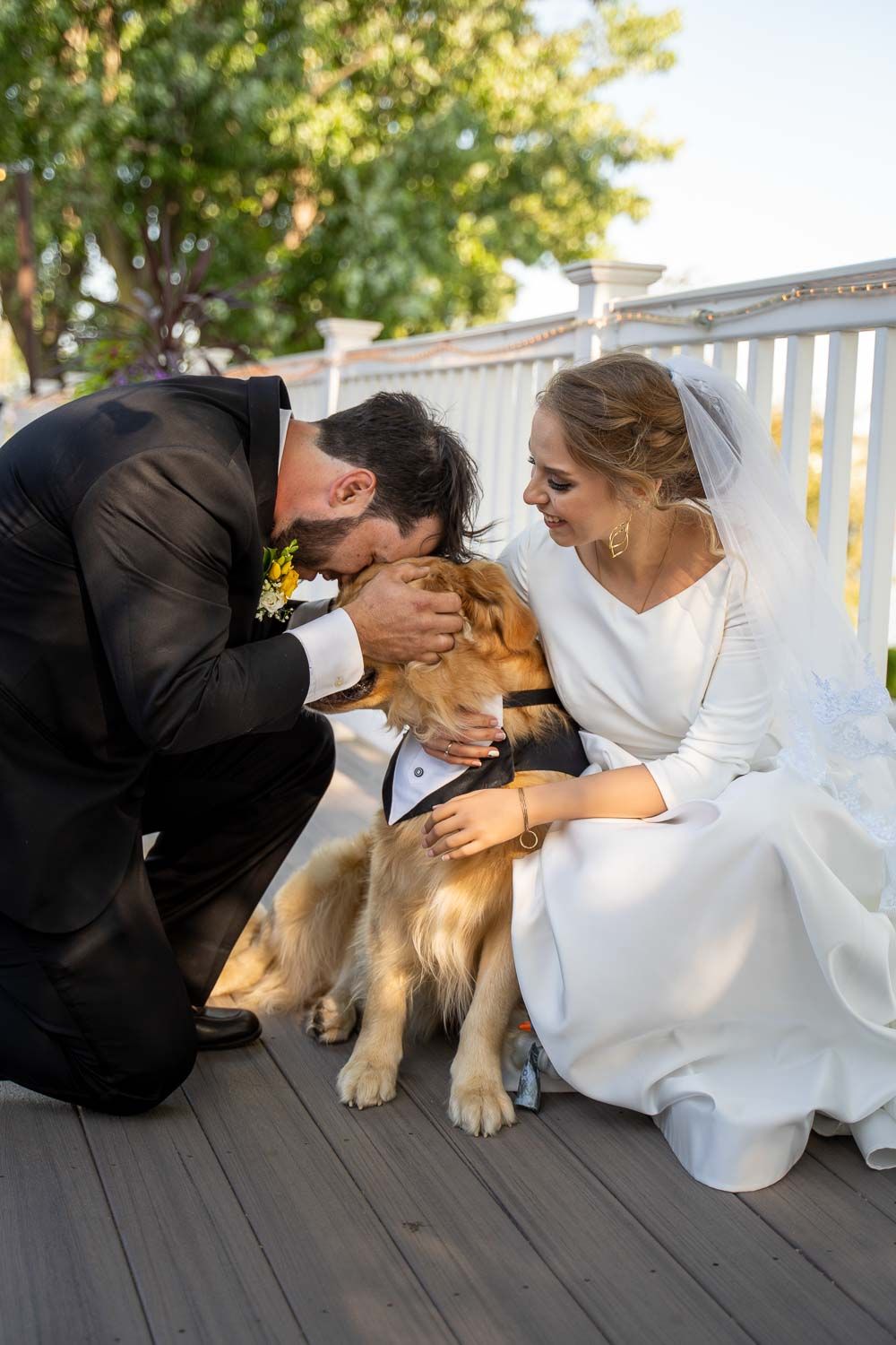 Bride and groom kneel on deck kissing their golden retriever dressed in a tiny tuxedo.