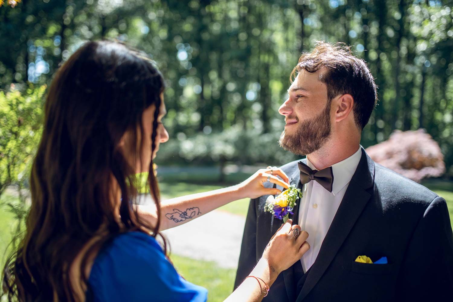 Woman with tattoo pinning floral boutonniere on smiling bearded groom in black tuxedo outdoors.