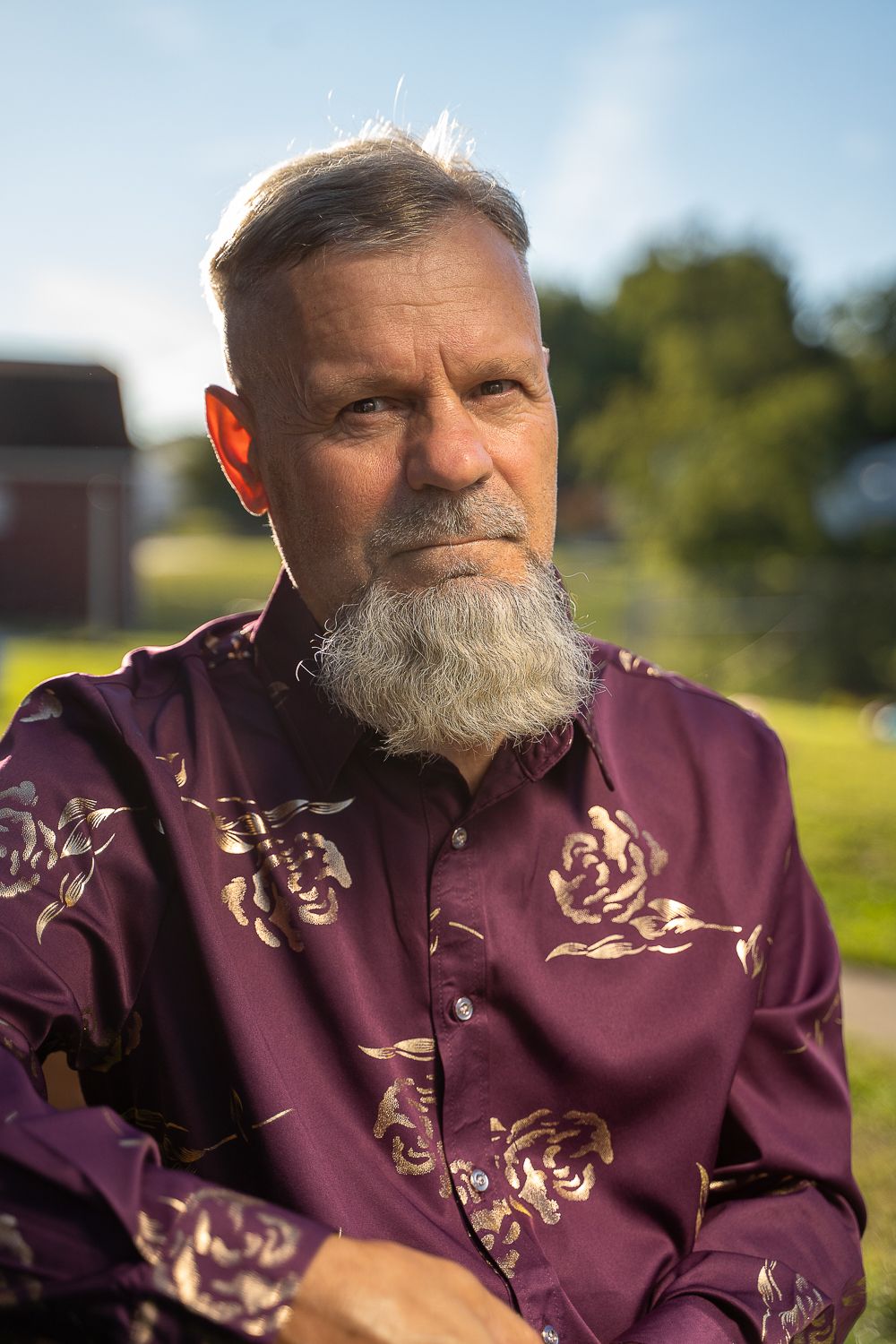Older man with gray beard wearing a purple floral shirt sitting outdoors.