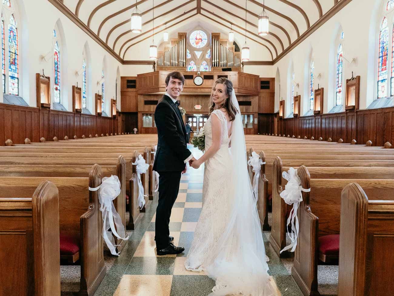 Smiling newlyweds pause in the aisle of a bright church with wooden pews and a pipe organ.