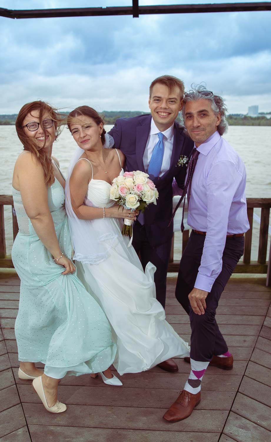 Bride with pink bouquet poses with groom and two guests on windy waterfront deck after wedding.