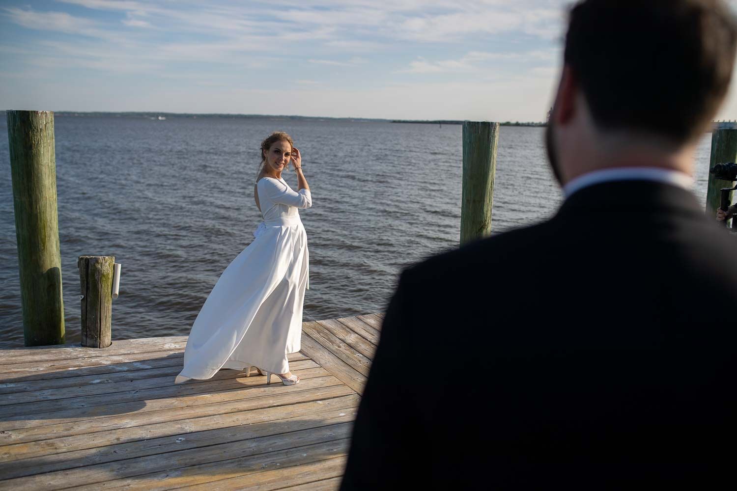 Bride in elegant white gown stands on waterfront dock as groom approaches during wedding portraits.