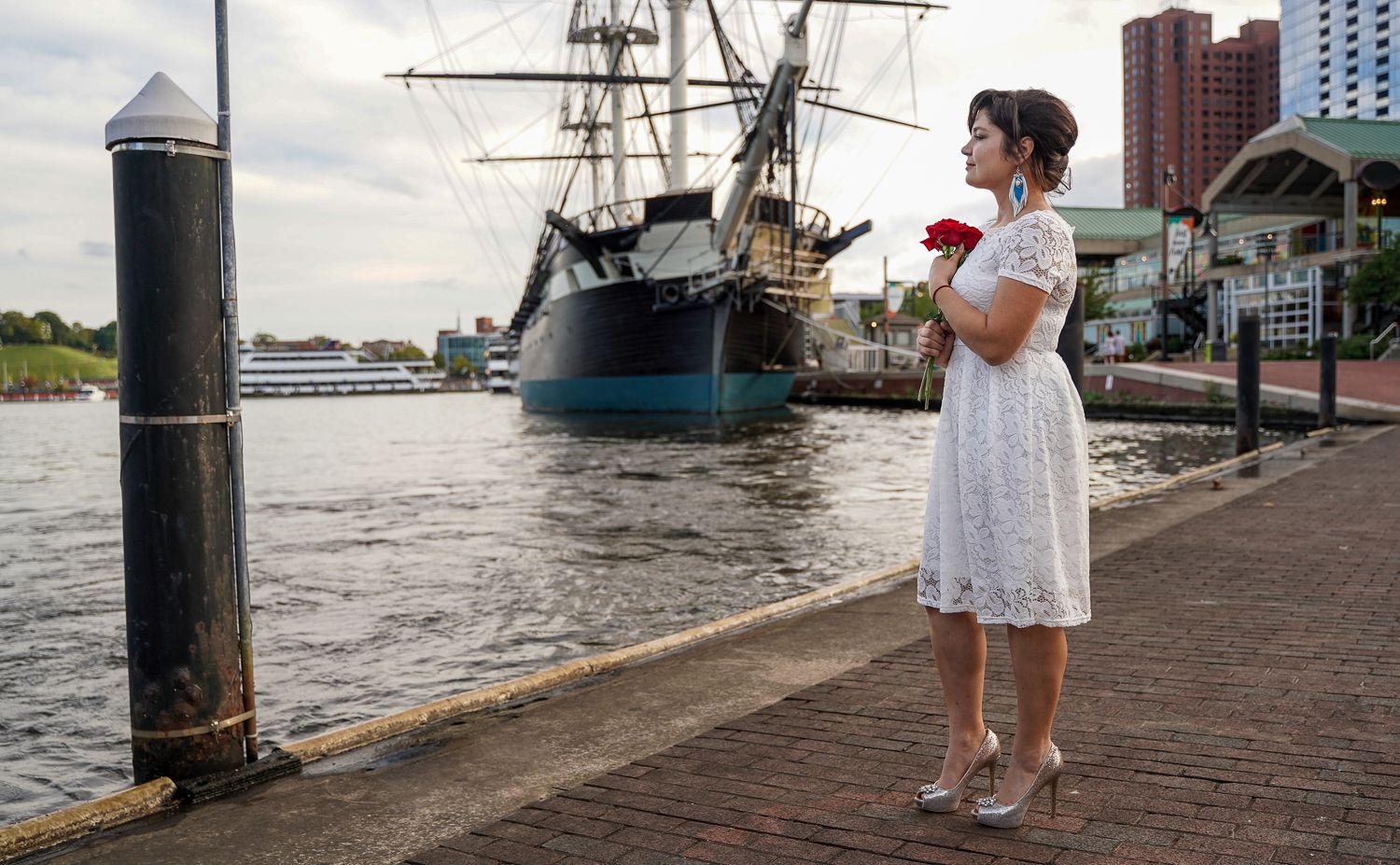 Bride in white dress holding red rose stands at harbor dock beside tall ship.