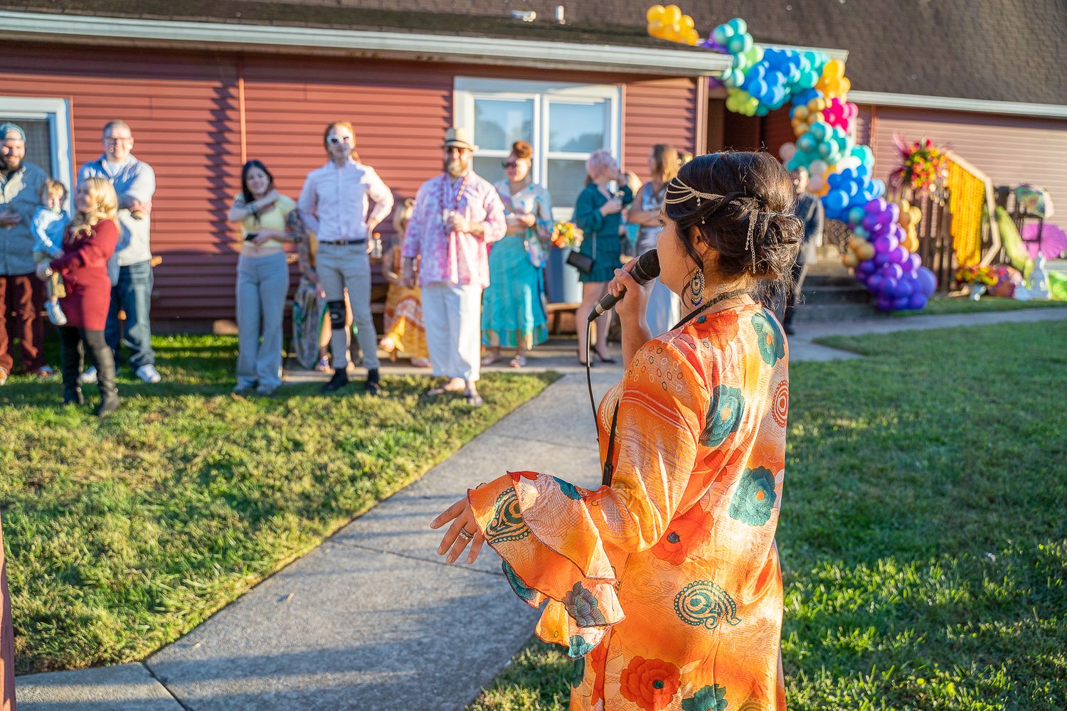 Woman in orange floral dress speaking into microphone at outdoor party with guests watching.
