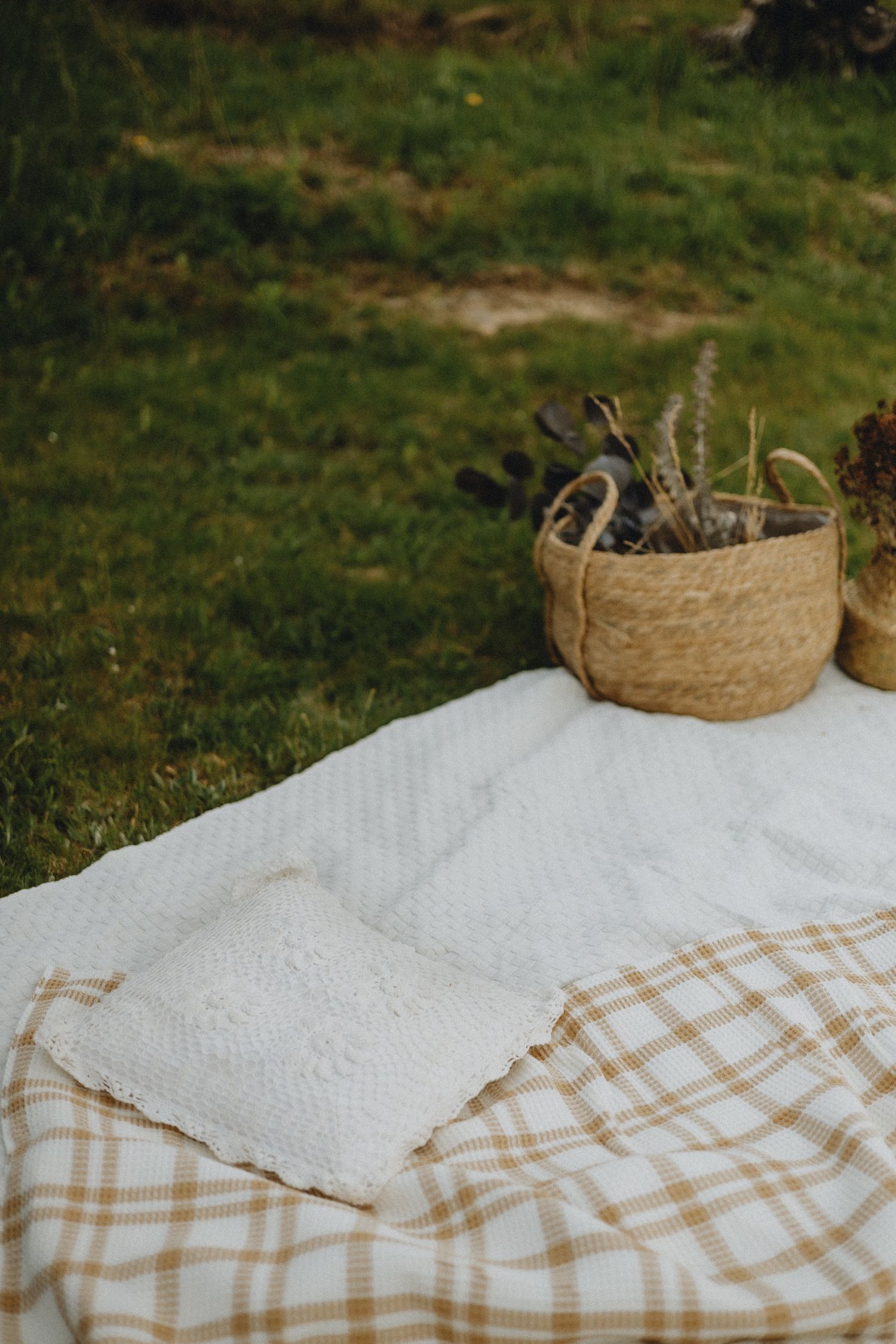 Woven basket with plants sits on checkered blanket and white linen against green grass background.