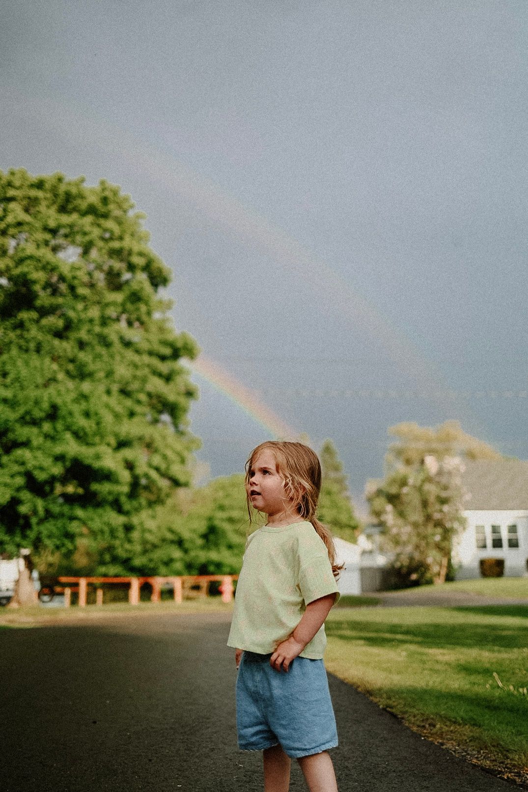 Double rainbow appears in sky behind child in green shirt and denim shorts.