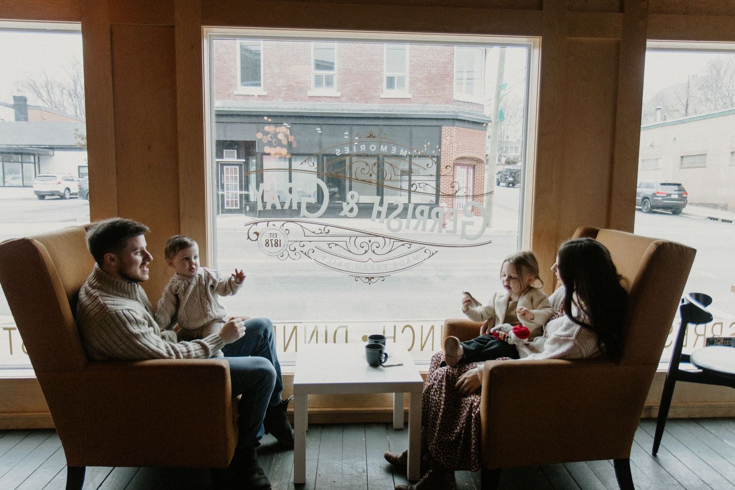 Two groups chatting in cozy armchairs by a large window overlooking a city street.