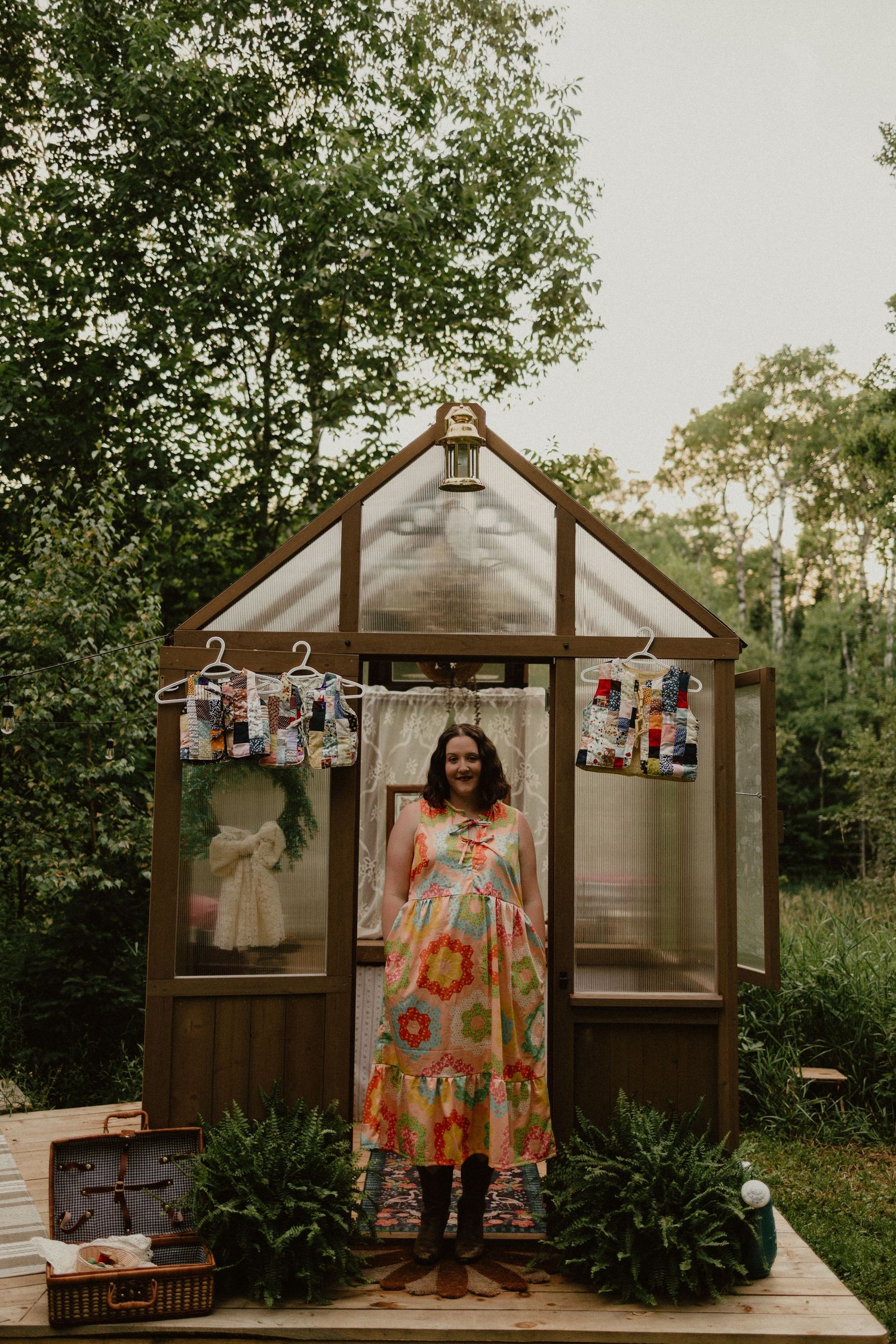 Someone stands in a floral dress outside a greenhouse decorated with photos on a wooden deck in a garden setting.