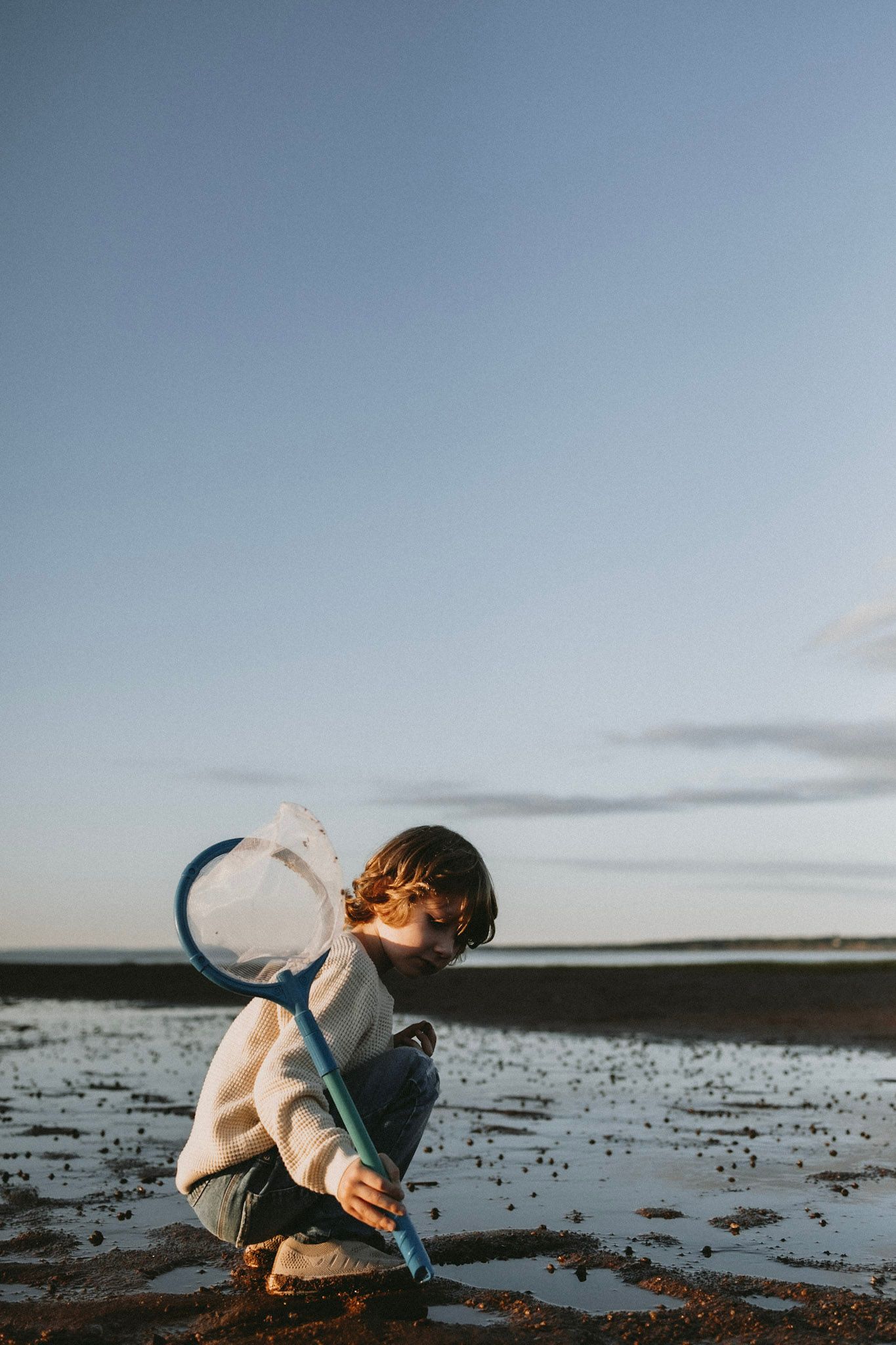 Child crouching in shallow water blowing bubbles with a wand against moody sky.