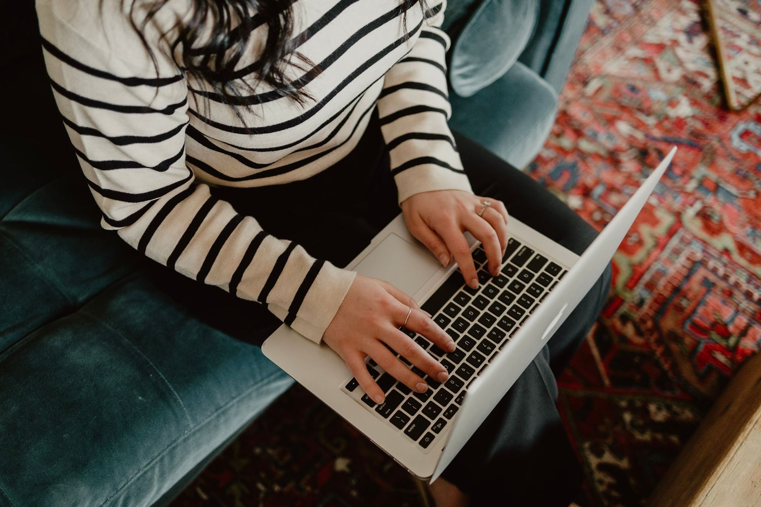 Hands in striped sweater typing on laptop keyboard while sitting on teal couch with patterned rug below.