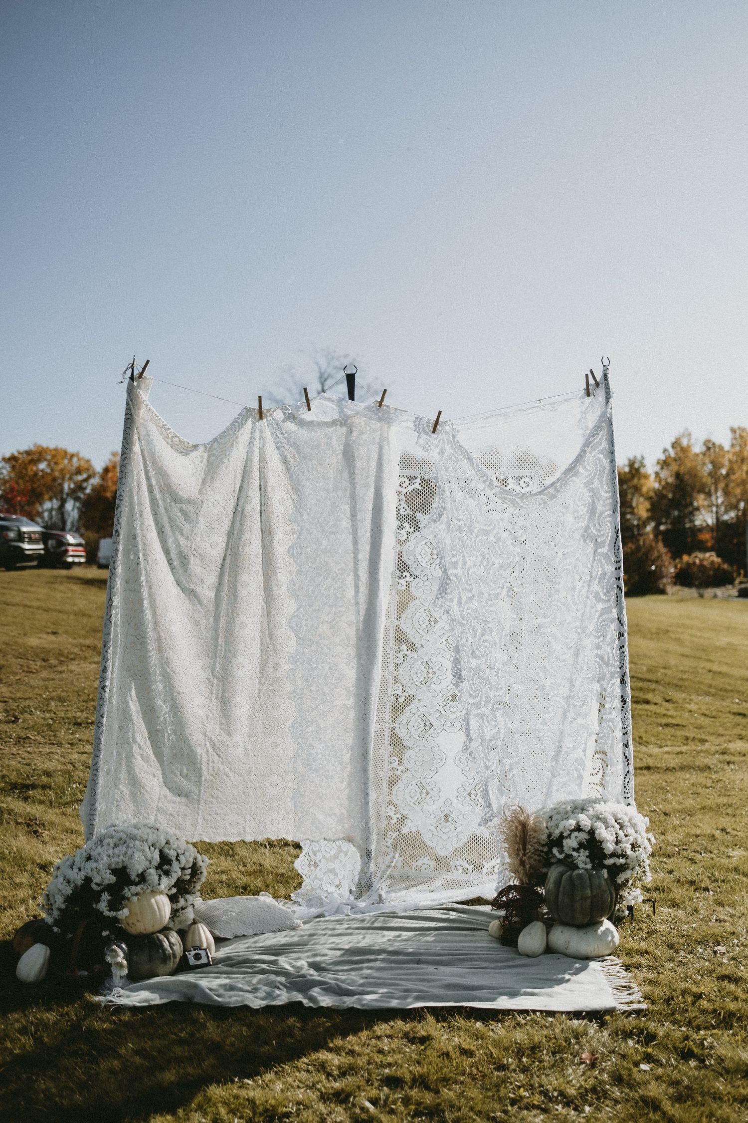 Rustic outdoor photo backdrop made of white lace curtains hanging on poles with floral arrangements at the base.