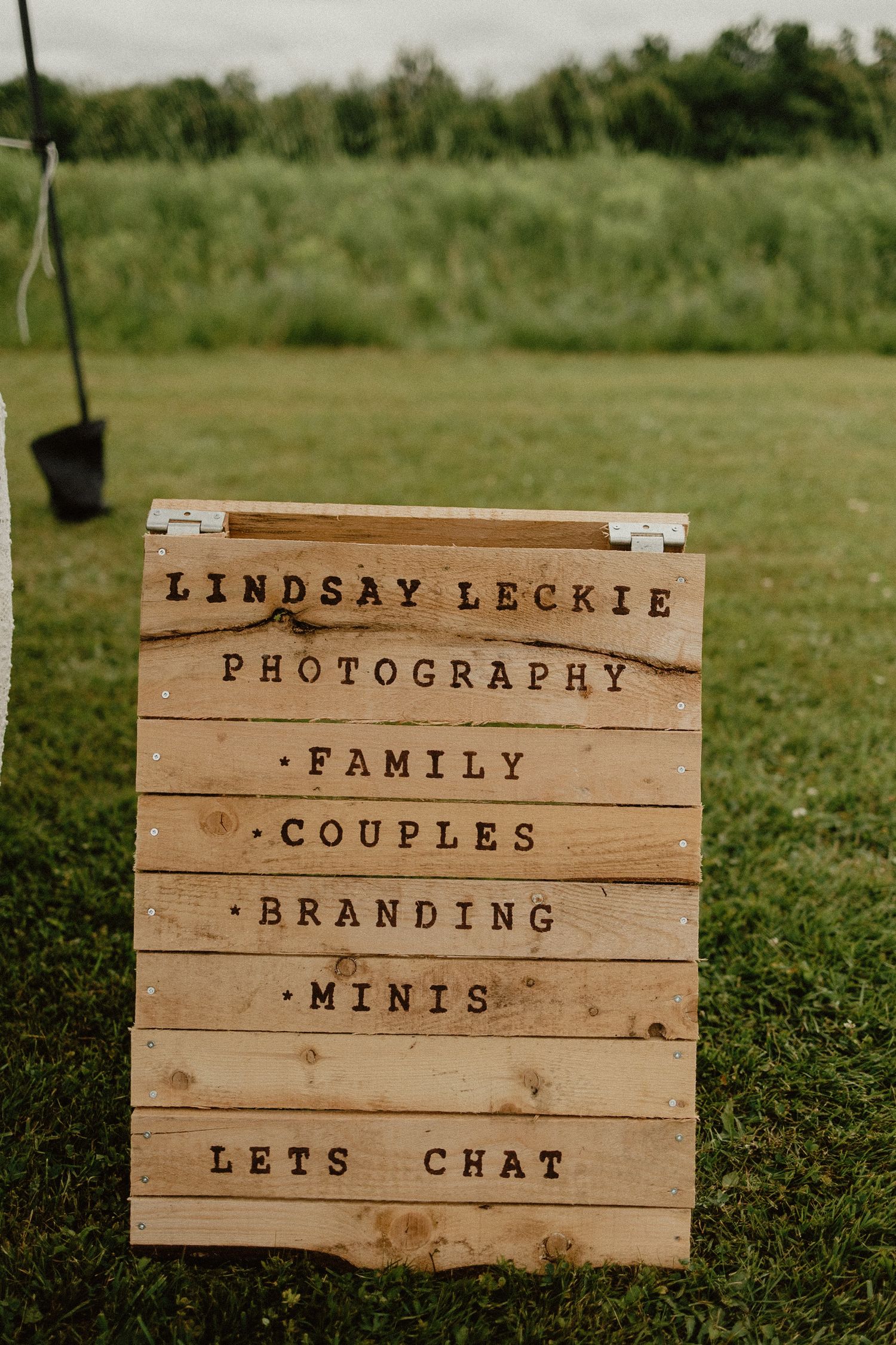 Rustic wooden sign displaying photography services including family, couples, branding and mini sessions on a grassy lawn.