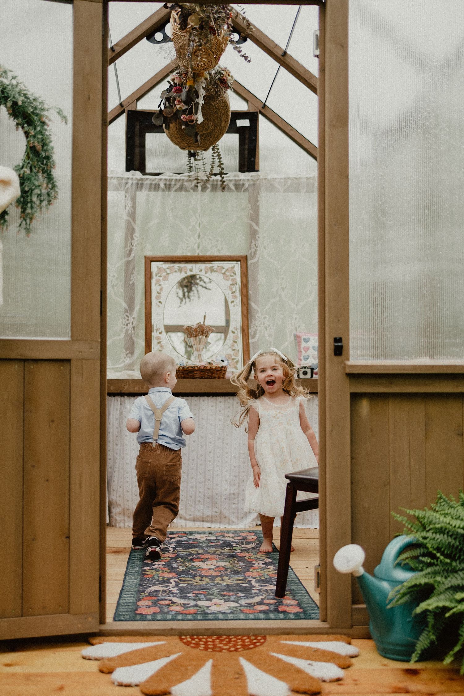 Two young children play and laugh together in a sunlit doorway of a cozy indoor greenhouse space with plants and decor.