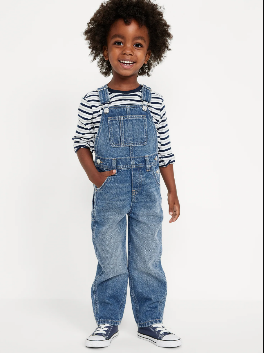 Child wearing striped shirt and denim overalls with blue sneakers.