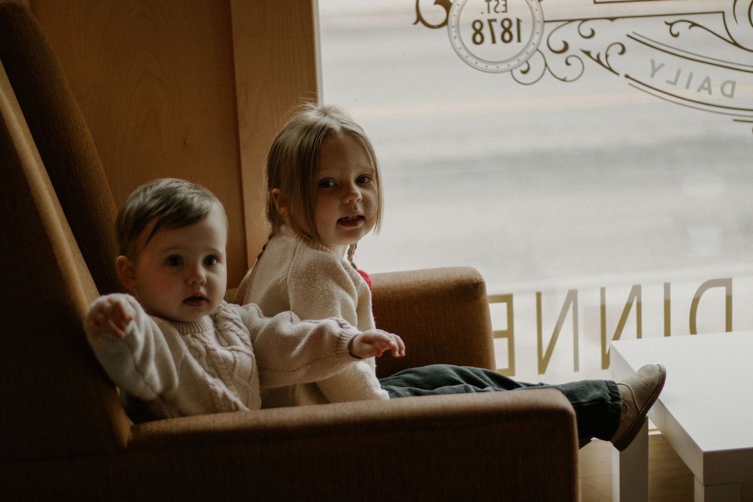 Two children sitting in brown leather chair by window.