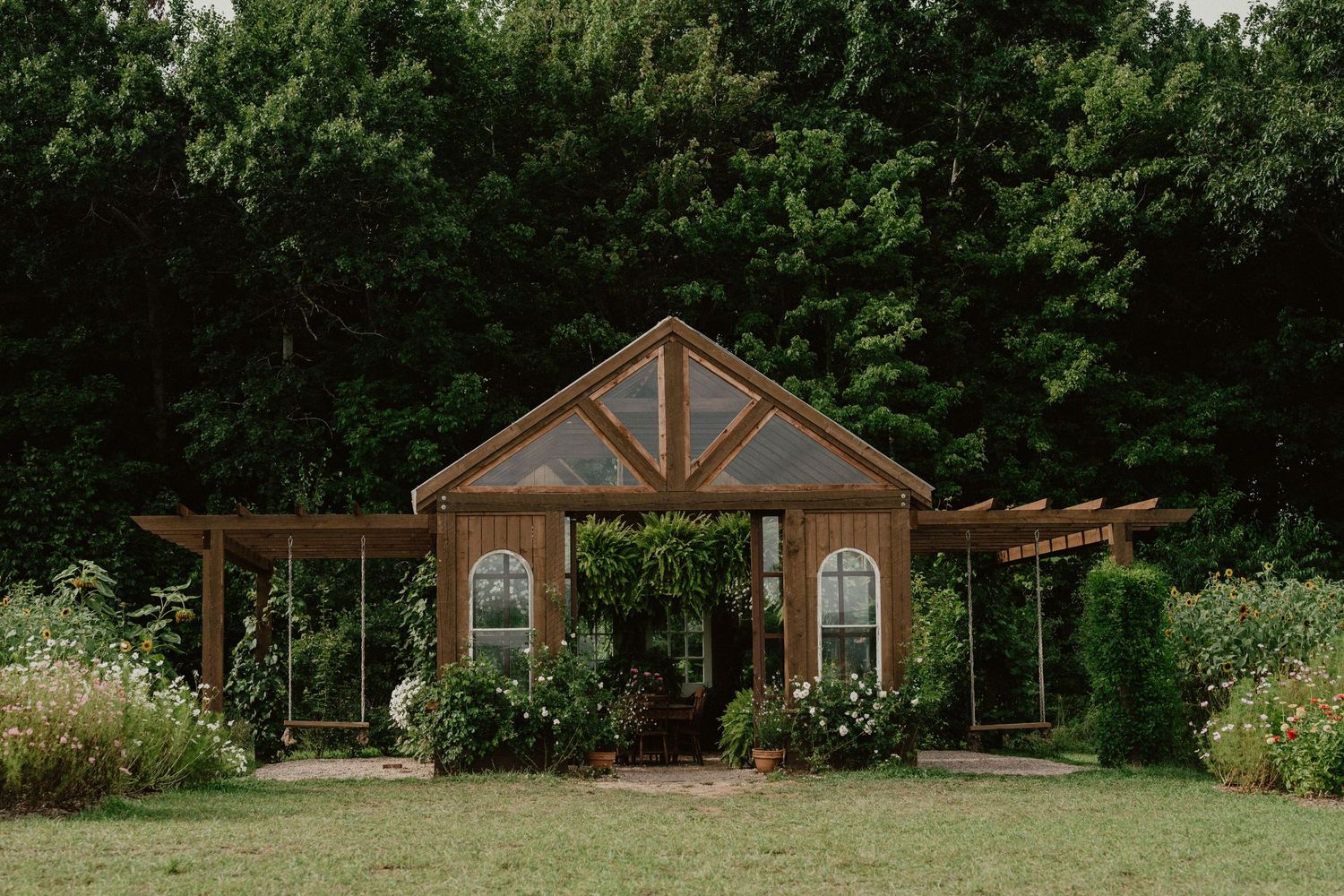 A rustic wooden wedding chapel with arched windows and pergolas sits nestled among lush greenery and gardens.