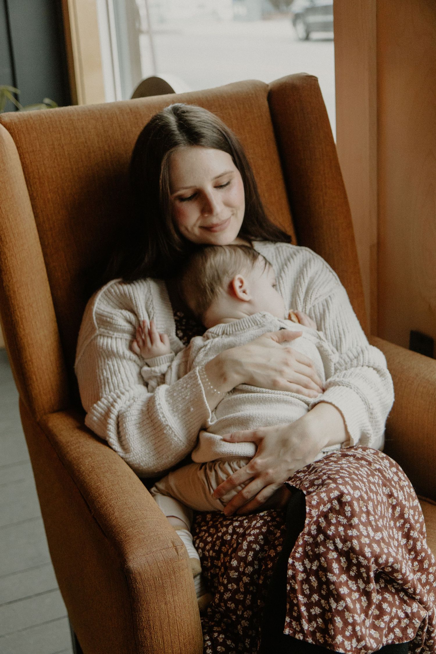 A tender moment between parent and baby in a brown armchair by the window.