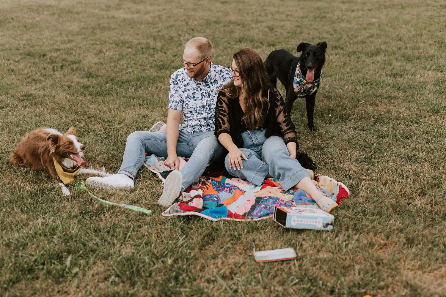 Couple having a picnic on blanket with their dogs in grassy field.