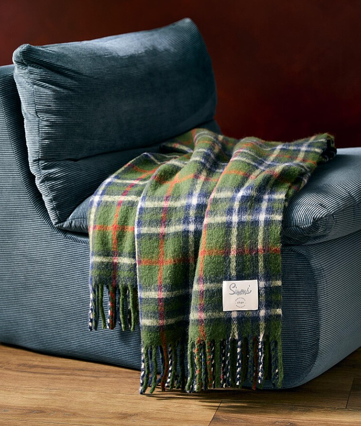 Plaid green and orange wool throw blanket with tassels displayed on a dark gray couch beside wooden floor.