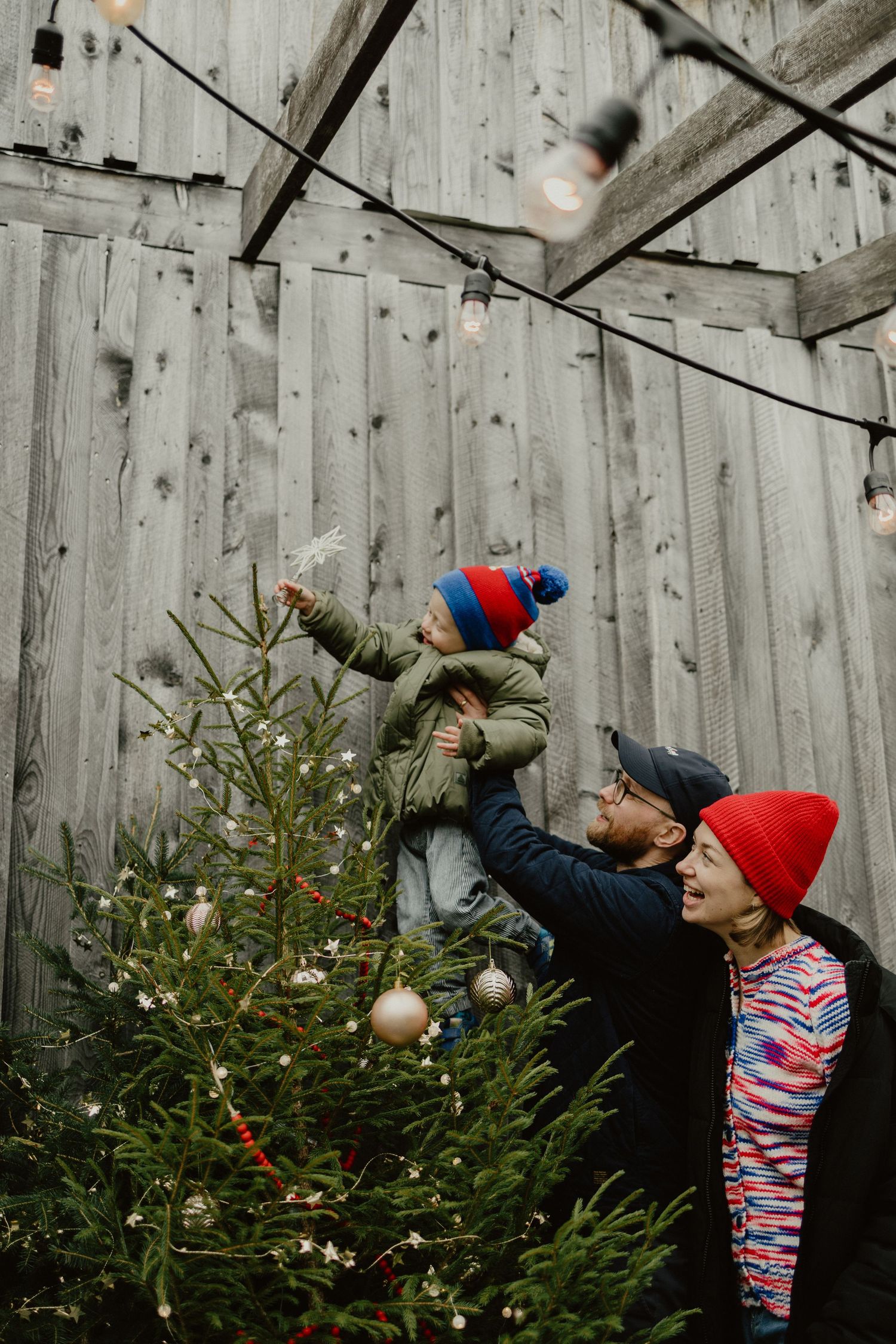 Family decorating a Christmas tree together outdoors by a rustic wooden barn wall during winter.