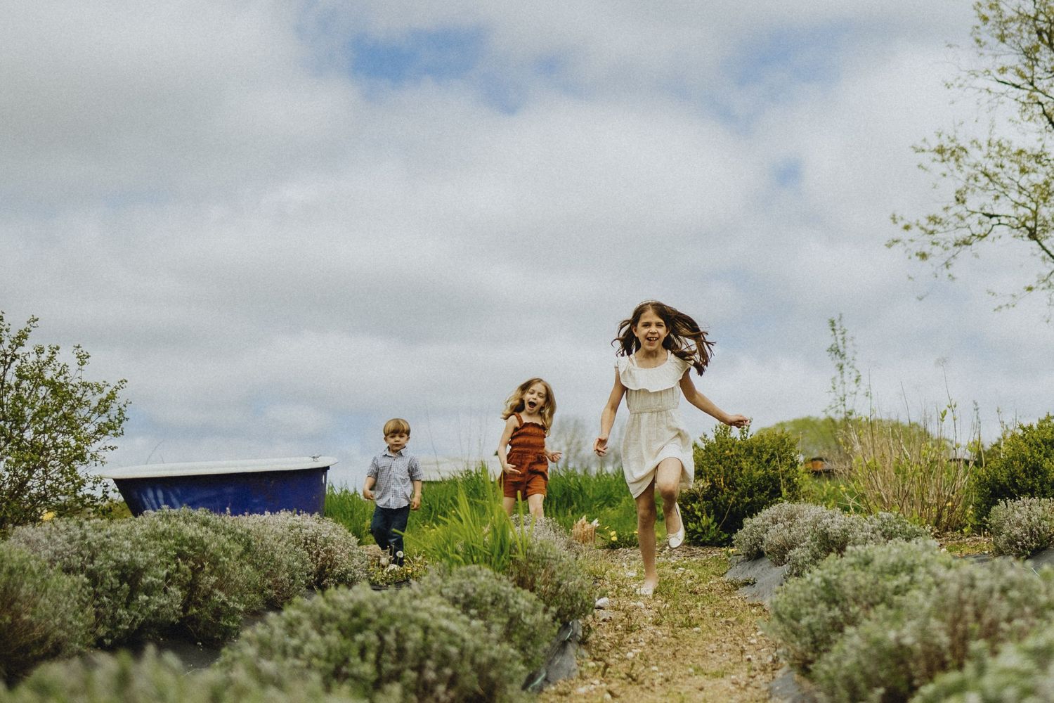 Children run through a wildflower field on a cloudy day with mountains in the background.