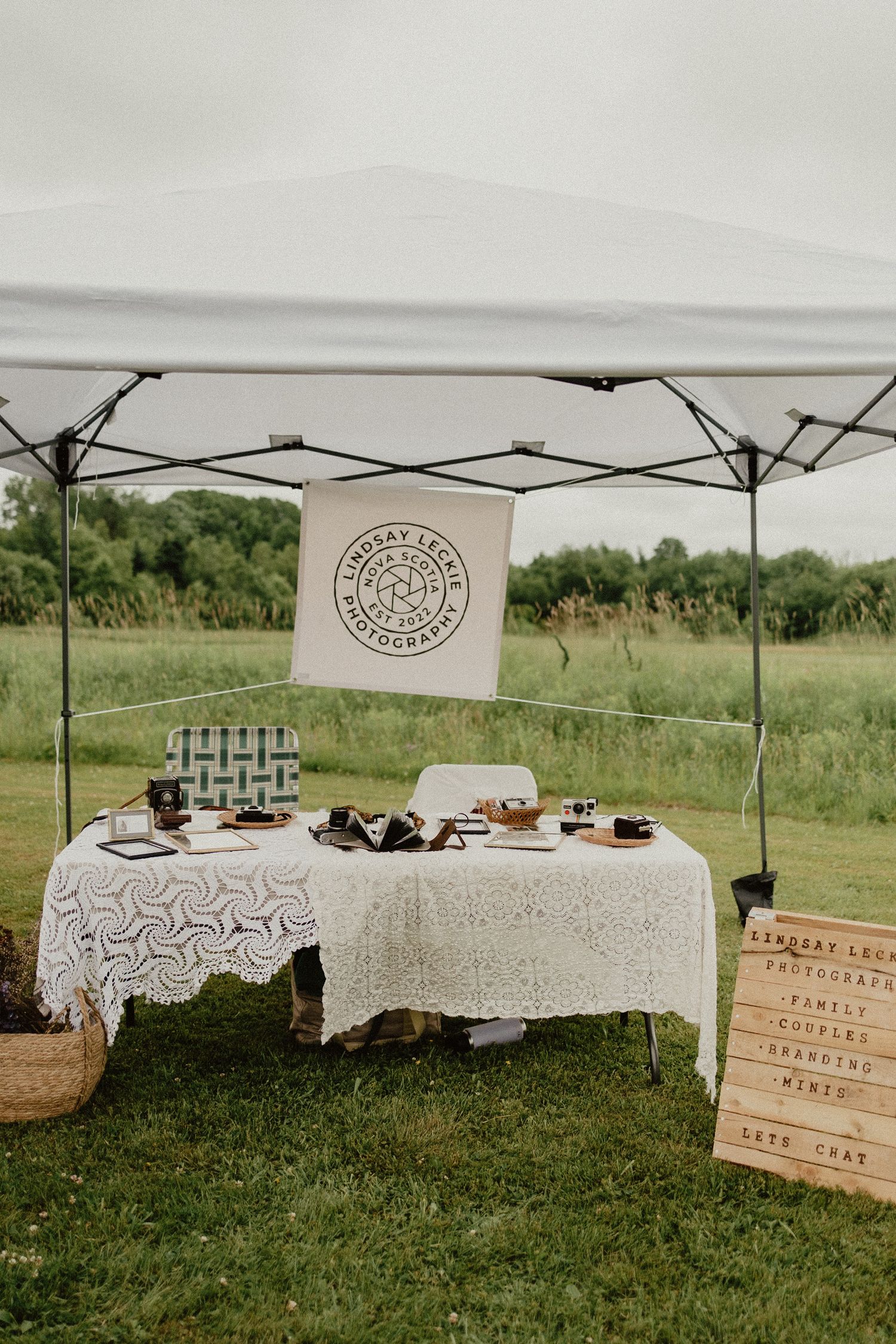 White pop-up tent with lace covered tables displaying food and drinks at an outdoor event setup in a grassy field area.