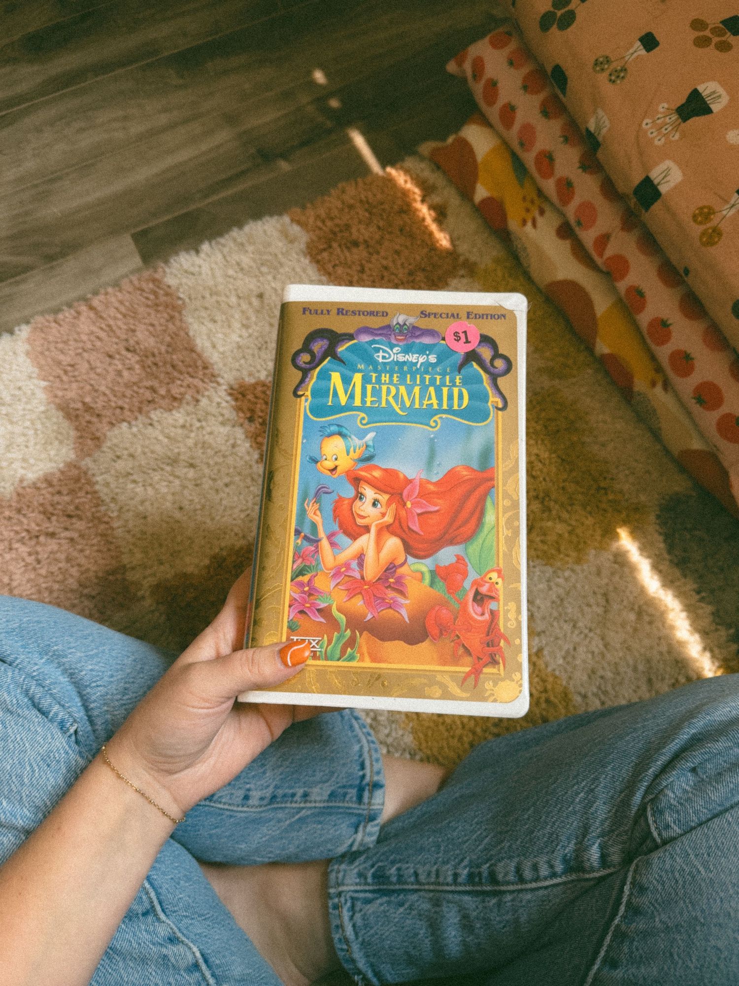 A person holding a vintage VHS copy of Disney's The Little Mermaid while sitting on a carpeted floor in sunlight.