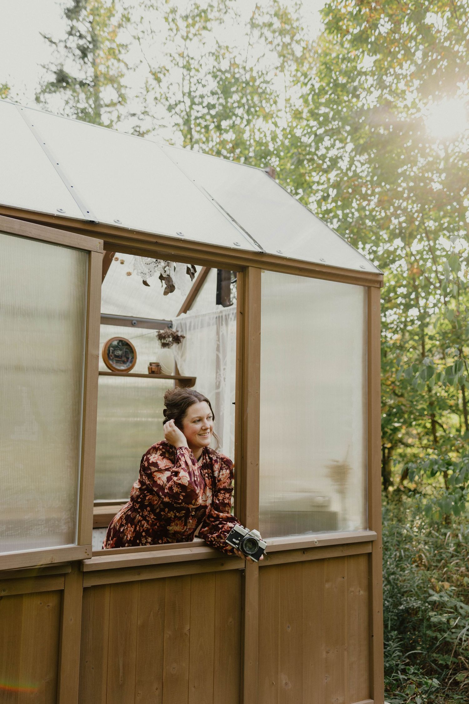 Person in brown jacket leans out window of rustic greenhouse structure with trees visible in background.