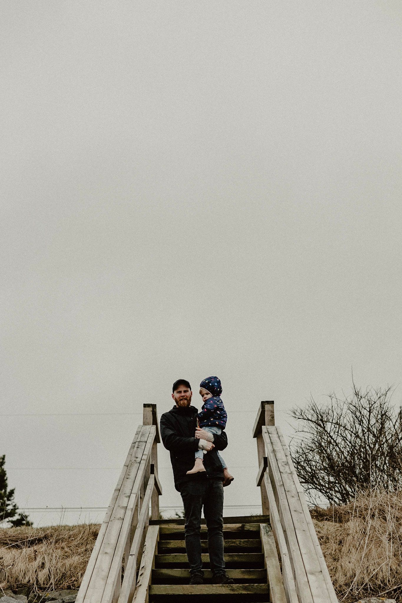 Two people stand together on wooden stairs overlooking a coastal landscape.