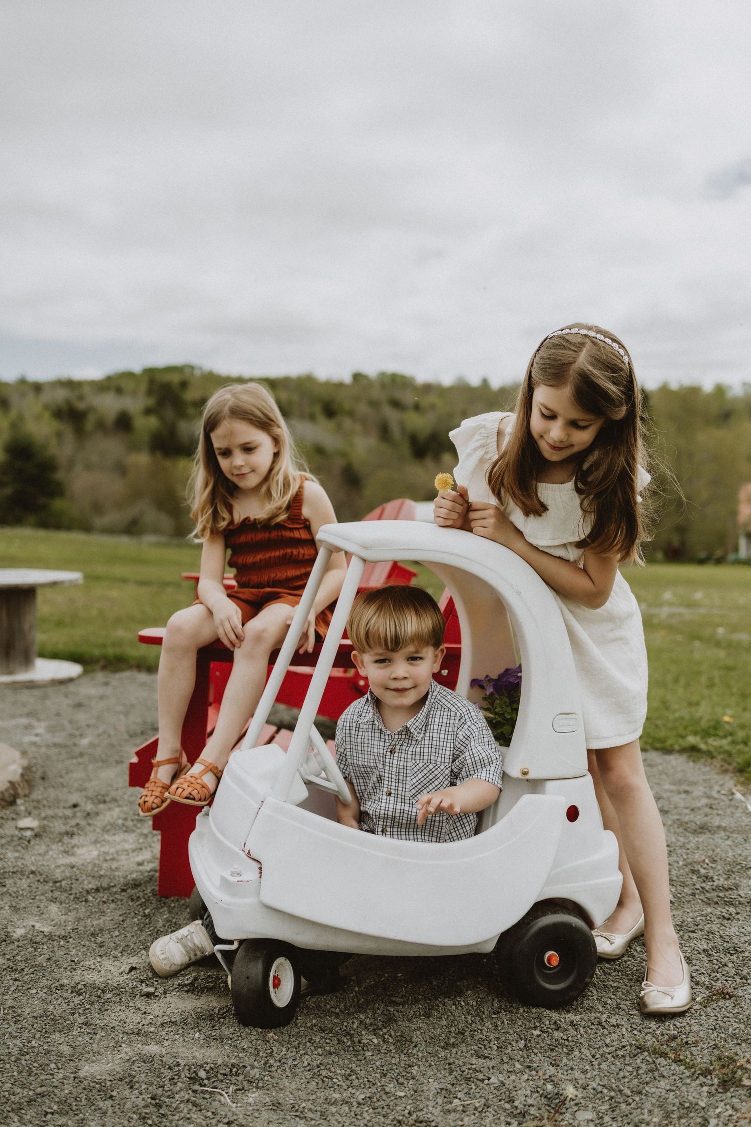Children playing with white toy car on gravel driveway during overcast day.