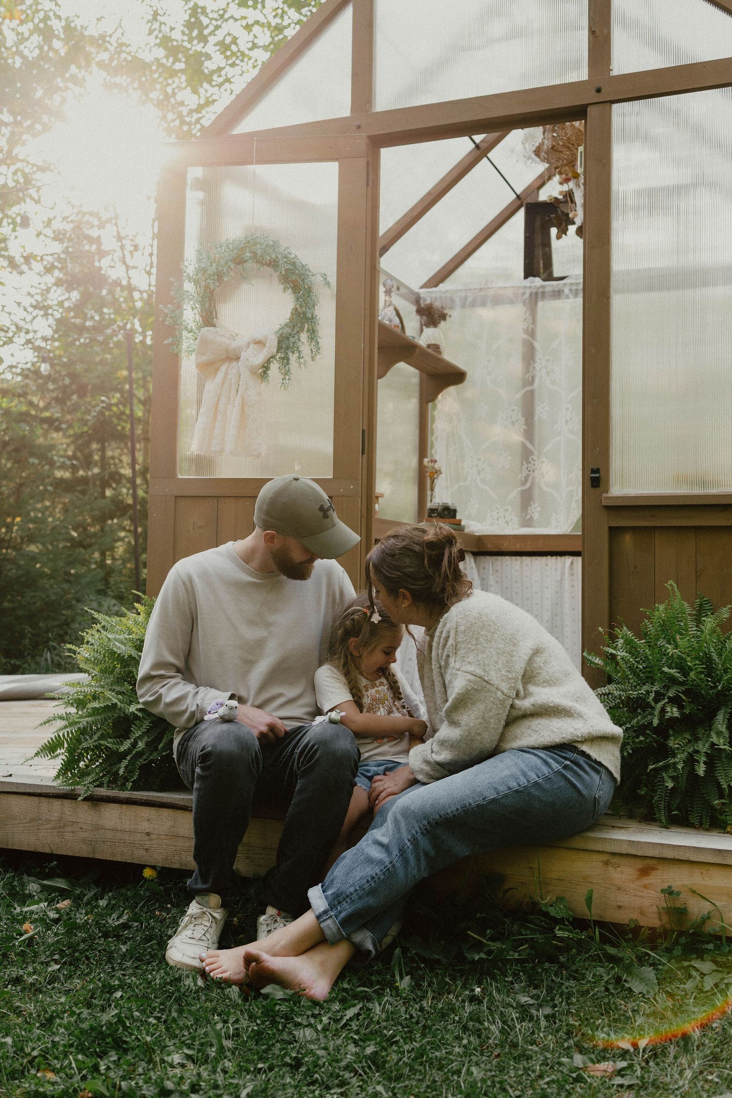 People sitting together on wooden steps outside a sunlit greenhouse with a wreath hanging at golden hour.