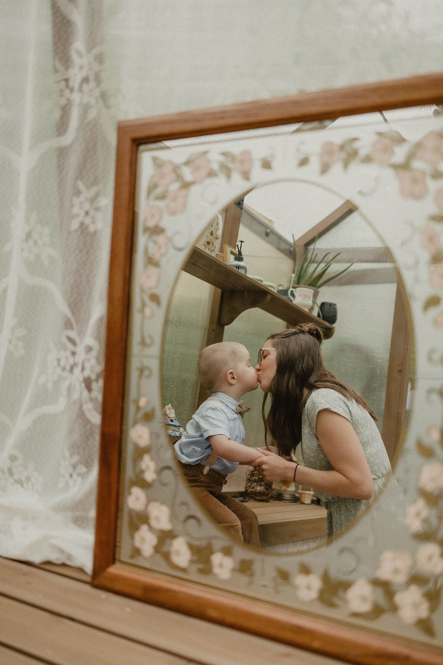 Mirror reflection shows mother and baby brushing teeth together in bathroom with floral wallpaper background.